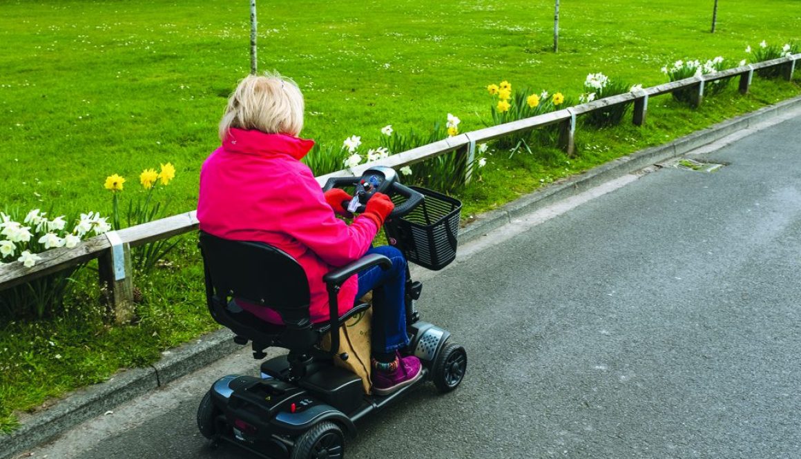 An elderly lady in a red coat enjoying the freedom of an electric mobility scooter on a quiet road with spring daffodils in bloom.