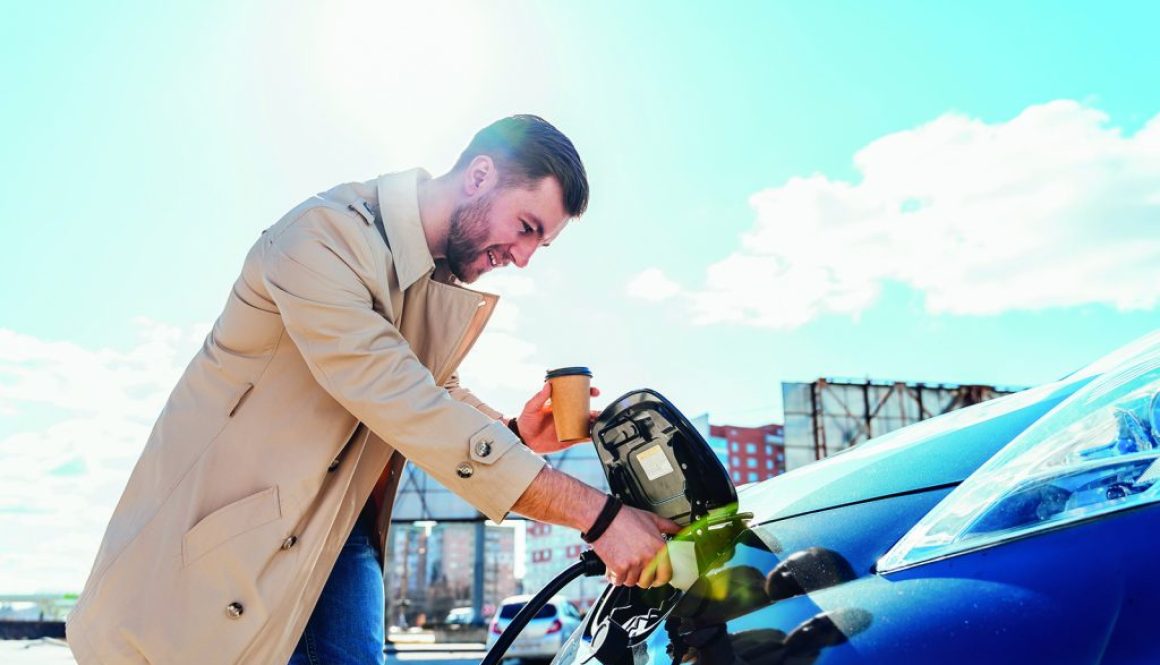 Stylish man with coffe cup in hand inserts plug into the electric car charging socket