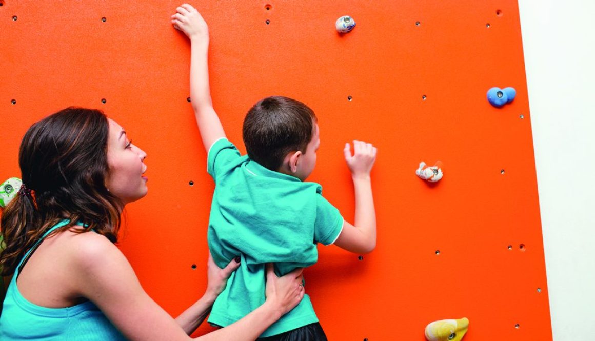 Mother helping her boy to climb on a wall in bouldering gym on playground